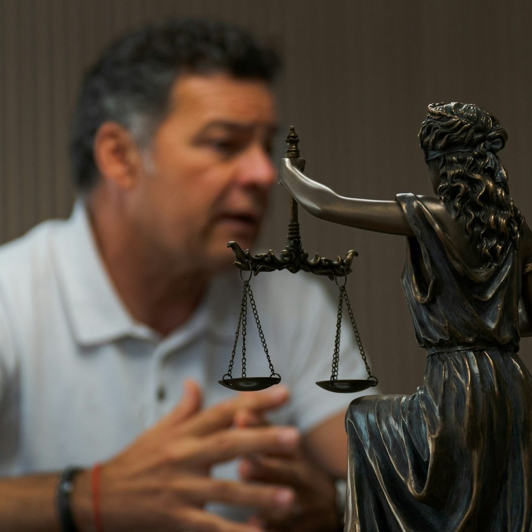 Pursue Justice A man in a white polo shirt sits behind a statue of lady justice.